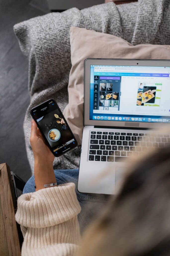 A woman multitasks, using a laptop and smartphone for design work, sitting on a sofa indoors.